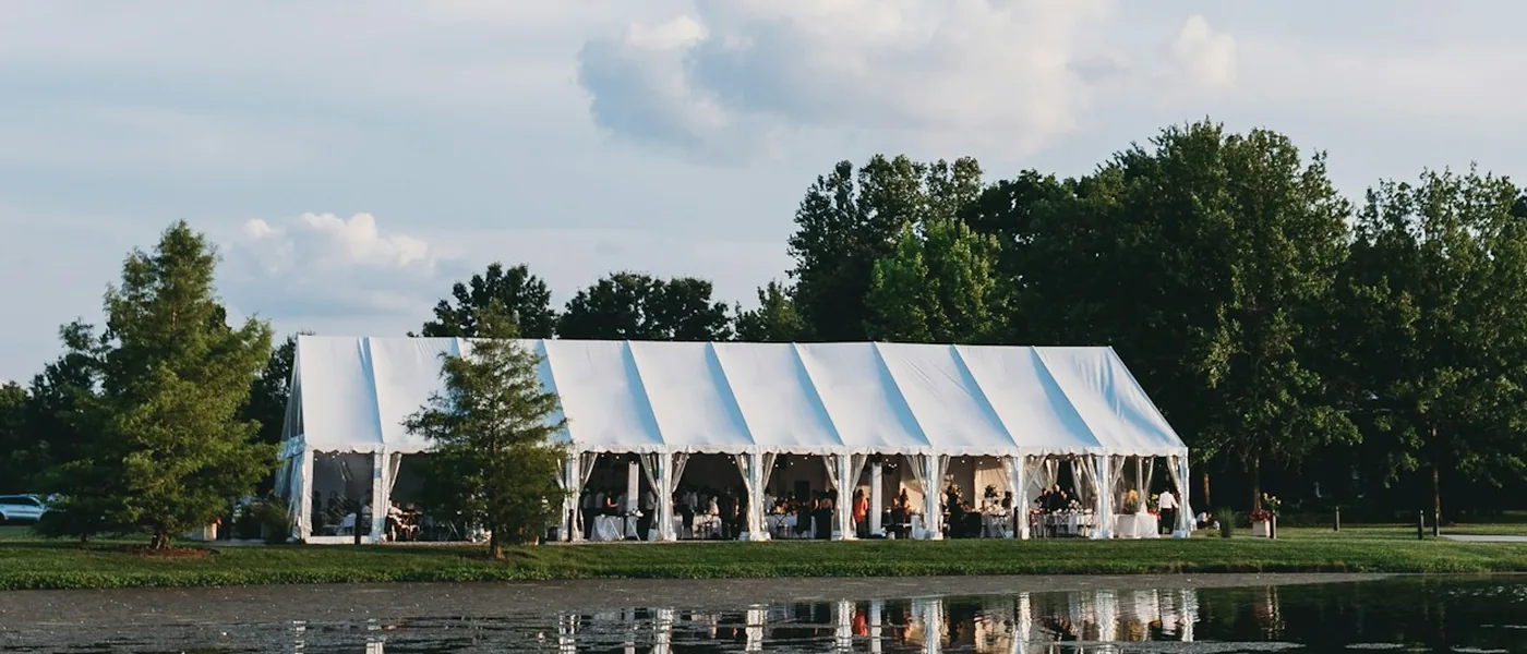 Large white frame tent with open sides set on a lawn beside a pond