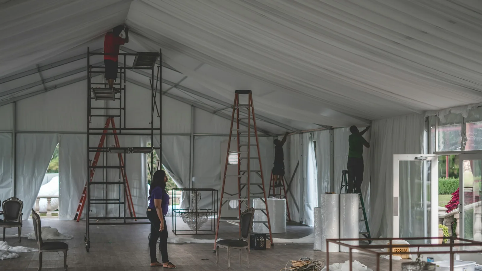 Crew on ladders and scaffolding finishing the interior of a large white frame tent structure